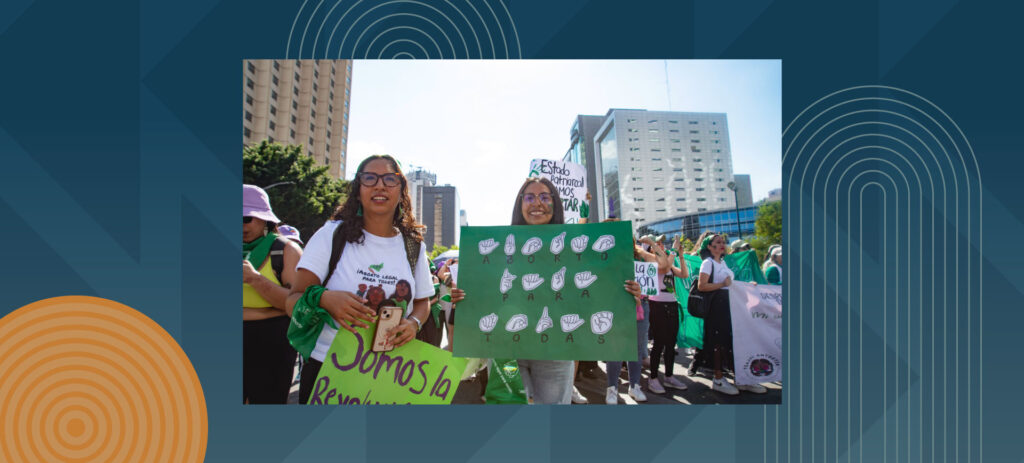 Two young women with glasses holding green protest signs at an outdoor demonstration in an urban setting, surrounded by other protesters wearing green and holding banners, with tall buildings visible in the background