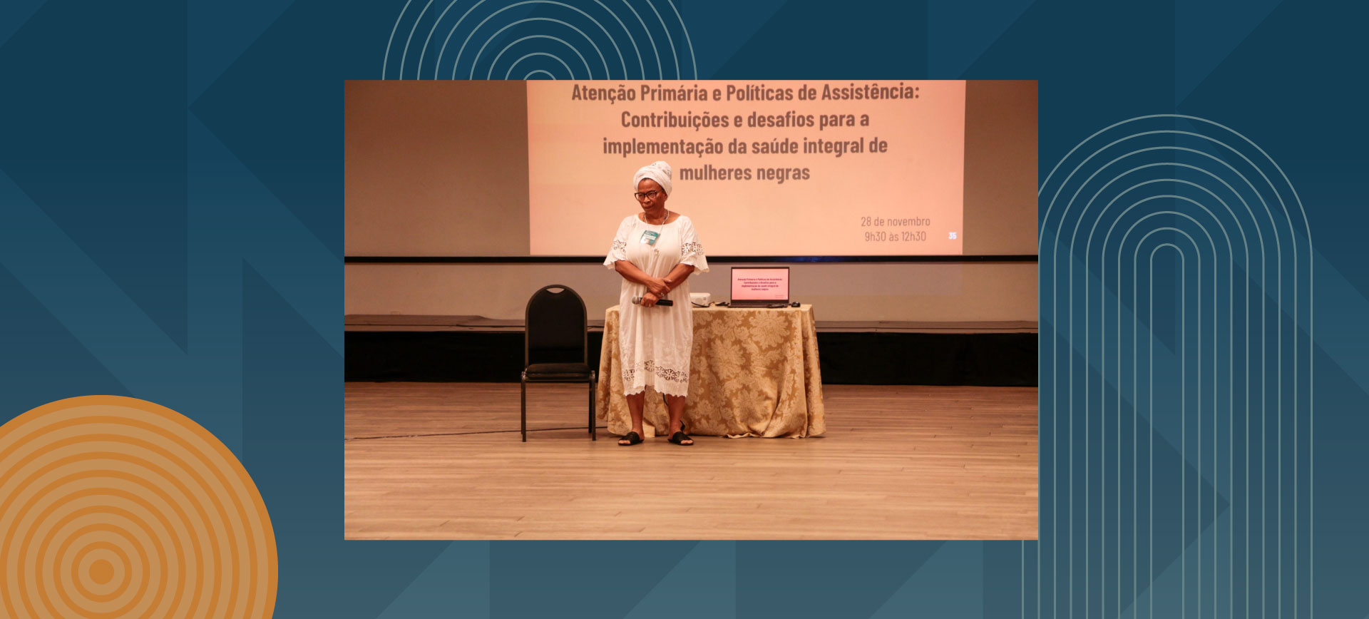 Black woman in white traditional dress and headwrap standing at a presentation table in an auditorium, with Portuguese text on screen about primary care and assistance policies for comprehensive health of Black women