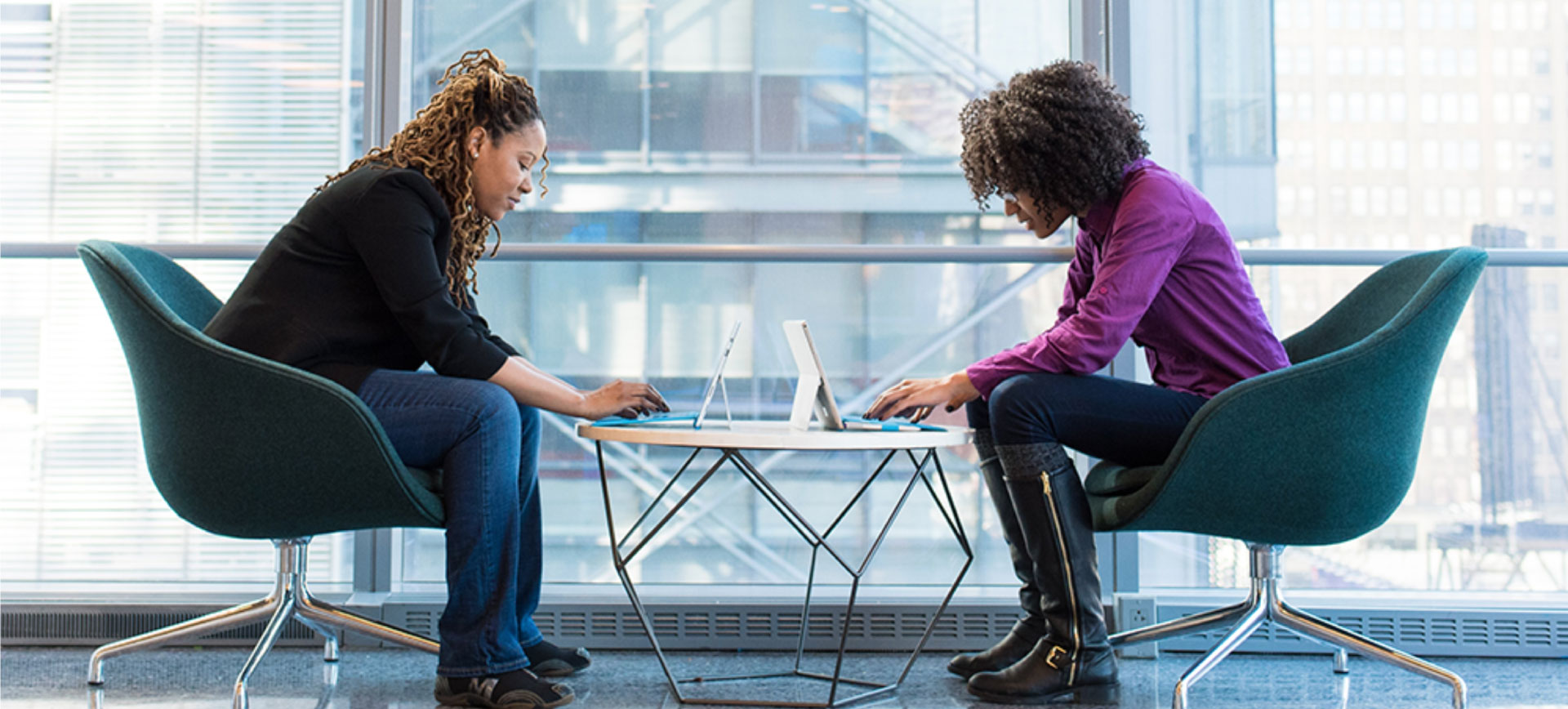 Two professional women working on laptops face each other in a bright, modern office with large glass windows.