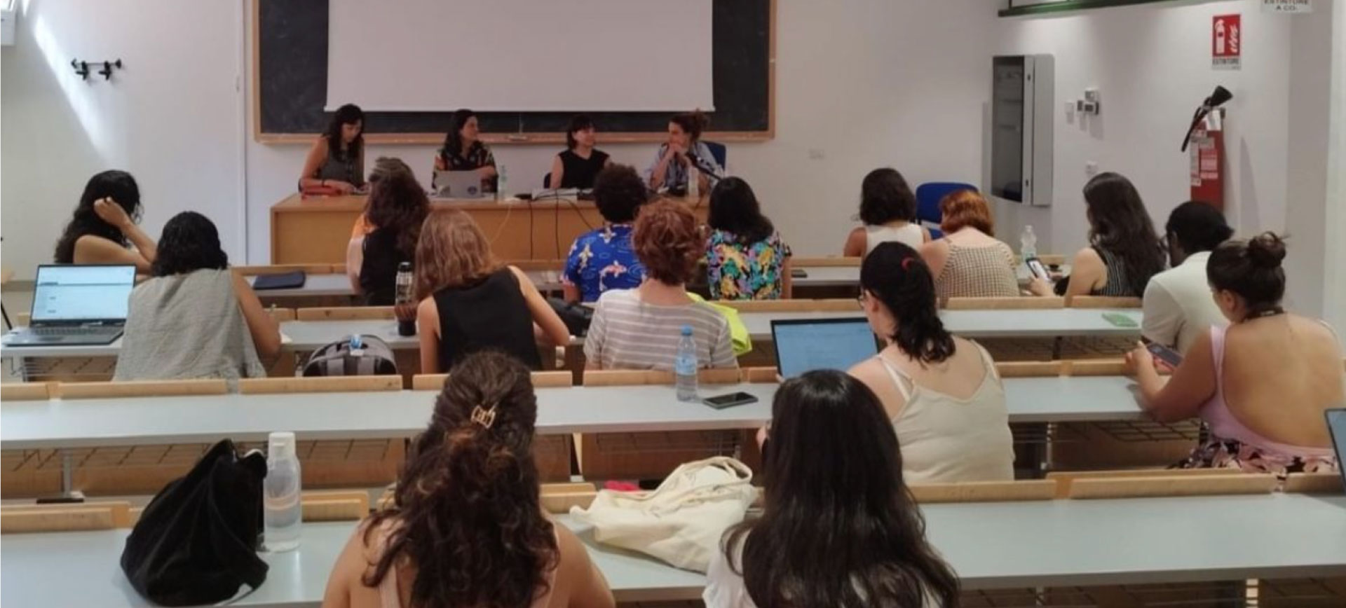 Four women panelists seated at conference table in front of audience in university lecture hall with tiered wooden seating, large screen backdrop, and attendees taking notes on laptops
