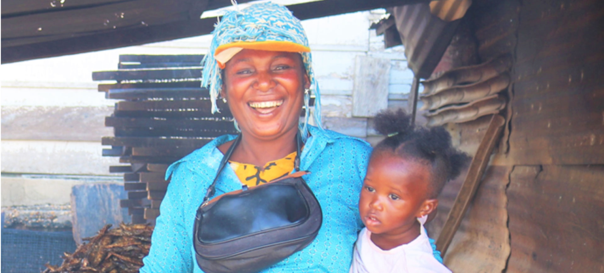 Smiling African woman in bright blue traditional dress and headwrap holding black bag, standing with young child in white shirt outside rural home with wooden structures
