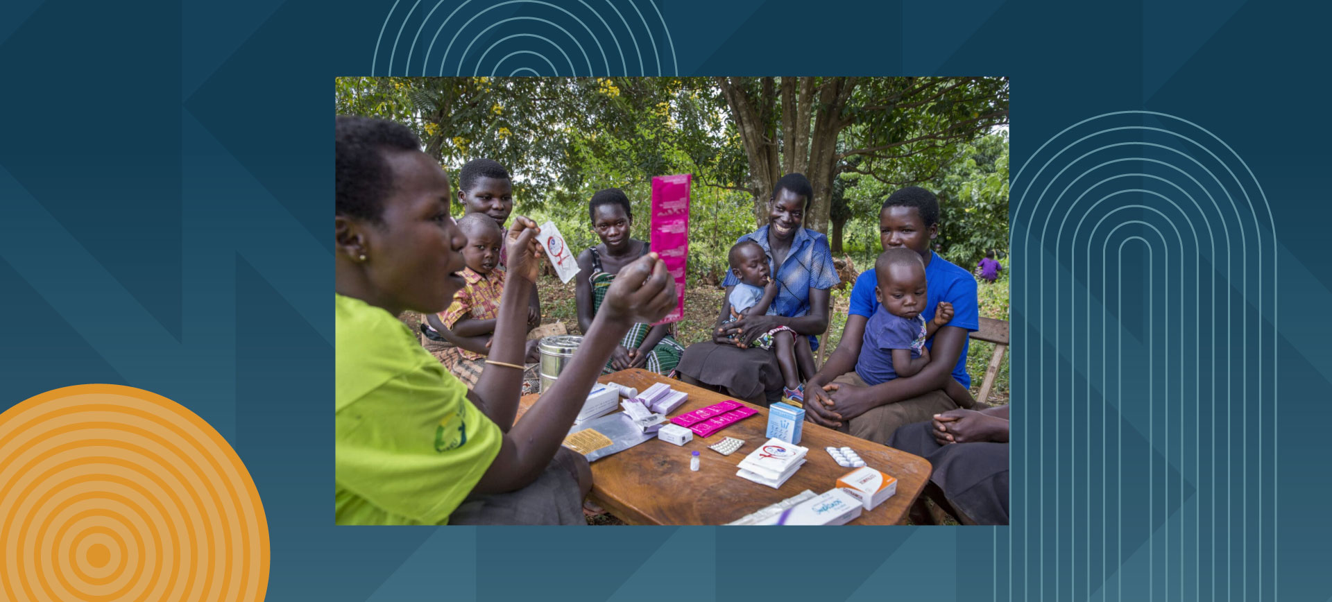 African healthcare worker in bright green shirt demonstrates medical supplies to group of mothers with children gathered around wooden table under trees, with various medical packages and materials displayed